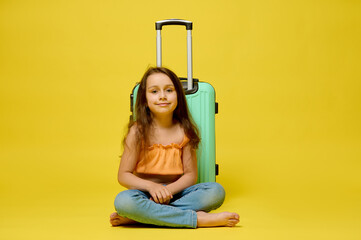 Caucasian cute traveler child girl with a suitcase, sitting over yellow studio background, smiling, looking at camera.