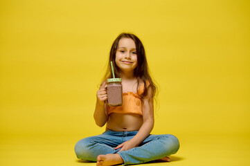 Smiling little girl drinks a cocktail from a glass bottle with a straw, isolated over yellow studio...