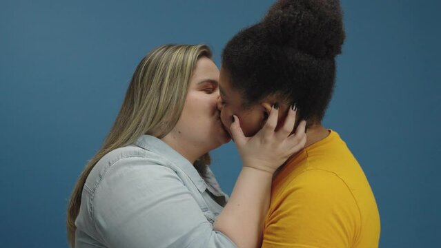 Sweet and tender kiss between two women in love shown of affection on isolated blue background studio. Free love to all sexual orientation. Gay young relationship.
