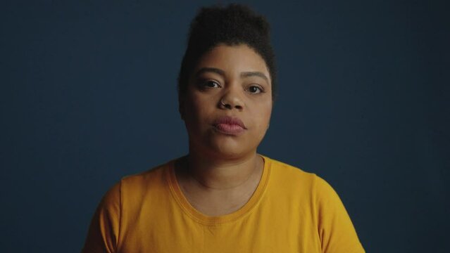 Serious young black woman looks into the camera with a serious expression. A close-up portrait of a Girl with a tired or sad look on a dark blue background. Dramatic cinematic female portrait.
