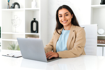 Female business photo. Elegant successful indian or arabian business woman, company employee sitting at workplace in the office, using laptop, working on a project, looks at camera, smiling friendly