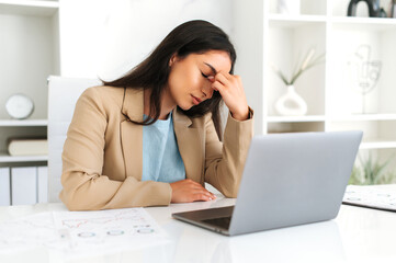 Overworked arabian or indian business woman, company ceo, corporate manager, sitting at workplace, massaging her nosebridge with eyes closed, feeling headache, migraine, stress, need rest