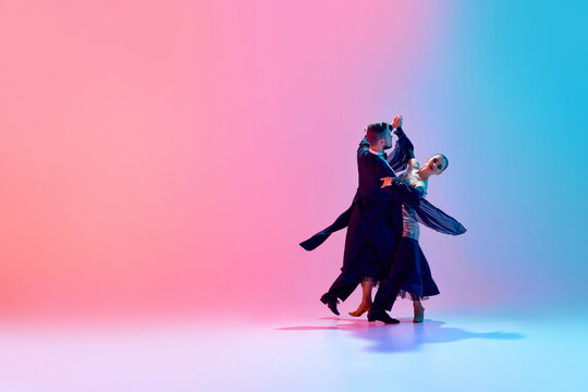 Young man and woman, talented ballroom dancers in motion, dancing in black costumes against gradient pink blue background in neon light. Concept of dance class, hobby, art, dance school, talent