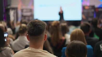 Back of a man's head in the foreground at a lecture. Engrossed young adults attend startup conference, woman speaker blurred in background with presentation. Casual dressed youth at business event
