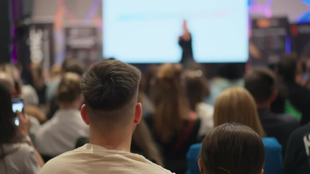 Back of a man's head in the foreground at a lecture. Engrossed young adults attend startup conference, woman speaker blurred in background with presentation. Casual dressed youth at business event