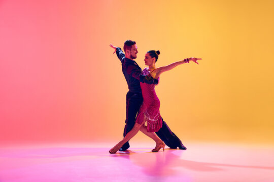 Passionate dancers. Artistic, talented young man and woman in motion in beautiful stage costumes dancing ballroom against gradient pink yellow background in neon light. Concept of dance class, art