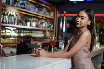Beautiful Asian woman wearing a white dress holding a glass of red wine drinking a cocktail at the bar counter, partying in a nightclub 