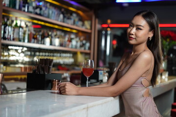 Beautiful Asian woman wearing a white dress holding a glass of red wine drinking a cocktail at the bar counter, partying in a nightclub 