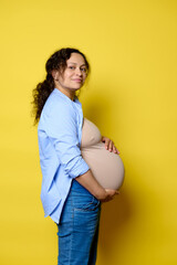 Happy pregnant woman putting hands on belly in last trimester of pregnancy, smiling at camera,...