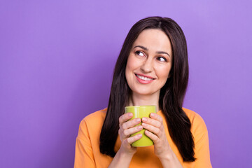 Photo of toothy beaming woman dressed orange t-shirt hold mug of coffee look empty space offer isolated on purple color background