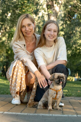 Mother and daughter squat down park paved road with dog enjoying walk. Smiling girl pets Shih Tzu spending quality time with parent in nature