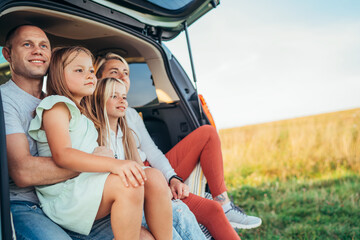 Portrait of Happy young couple with little daughters sitting inside car trunk during auto trop. They are smiling, laughing and chatting. Family values, traveling concept.