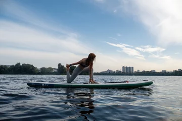 Poster Young woman balances on SUP board stretching legs to engage muscle groups. Lady relaxes gliding on river surface on paddleboard © SlavaStock