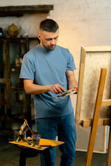 in an art workshop an artist in a blue T-shirt picks up paint on palette with a palette knife
