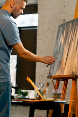 Fototapeta na wymiar in an art workshop an artist in a blue T-shirt makes broad strokes on the canvas with palette knife