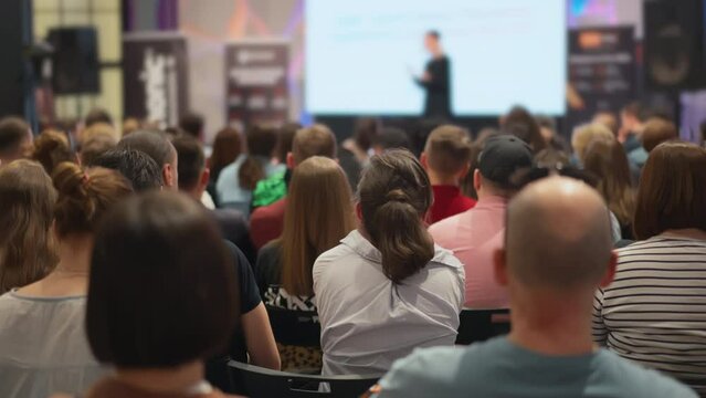 Audience of young entrepreneurs listens to a startup pitch, indistinct female speaker on stage with visuals. A scene of active young minds at an incubator event. Concept: youth initiatives, startup