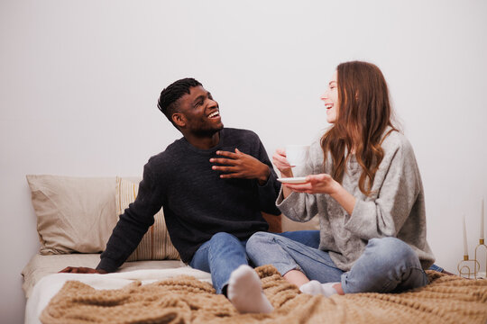 Cheerful multiethnic couple with coffee talking on bed at home 