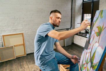 young artist in a blue t-shirt in an art studio working on painting while sitting