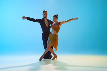 Elegant young man and woman, ballroom, Latin, samba dancers performing against gradient blue background in neon light. Concept of dance class, hobby, art, dance school, talent