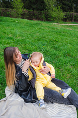 Fototapeta premium mother holds her son on her lap and sits on a blanket on a green lawn in a spring park