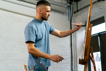 in an art workshop an artist in a blue T-shirt makes broad strokes on the canvas with wide and flat...
