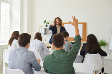 In the bustling school classroom full of students, a dedicated teenage schoolboy raises his hand, signaling active participation during a lesson. The teacher attentively guides the learning process.