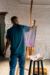 in an art workshop an artist in a blue T-shirt creates pink smudges with brush on canvas