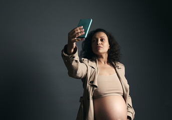 Beautiful mid adult pregnant woman with naked belly, in stylish beige trench, using mobile phone, over gray background