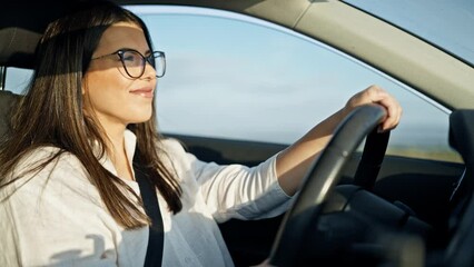 Young beautiful hispanic woman driving a car smiling wearing glasses on the road