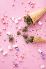 Obraz na płótnie Canvas Dry buds of medical marijuana lie on waffle ice cream cones on a pink background. There are candies and marshmellos around. Alternative medical cannabis treatment