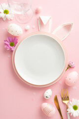 Chic Easter table layout concept. Vertical top view of jocular bunny ears protruding from plate, amidst adorned eggs, eating utensils, flute, flowers on pale pink foundation with empty space for copy