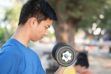 Asian plump young man lifts four-kilogram and eight-kilogram dumbbells to exercise and loose weight in an open courtyard in late afternoon, health care and recreational activity concept.