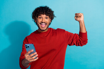 Photo of overjoyed hispanic guy wear red pullover raised fist up winning lottery auction with smartphone isolated on blue color background