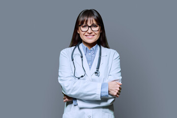 Confident friendly female doctor with crossed arms on gray background