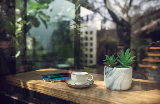 White coffee cup with mobile and note book, small tree in vase on wooden counter in cafe and outdoor garden background