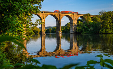 Panoramic view of railway viaduct with train crossing Ruhr river in Herdecke Germany. Historic brick construction with tall arches on the branch line from Hagen to Dortmund reflected by water surface. - obrazy, fototapety, plakaty