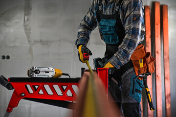 Fototapeta premium a worker in fitters and gloves with a tool belt measures wood using a tape measure and a right-angled triangle, blurred background