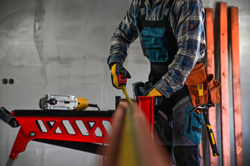 a worker in fitters and gloves with a tool belt measures wood using a tape measure and a...