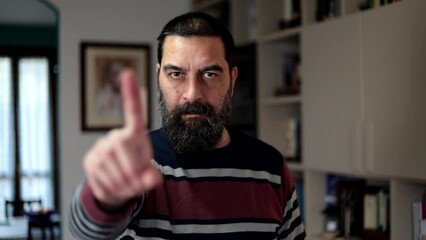 Bearded man standing with serious expression saying no with finger at home.
