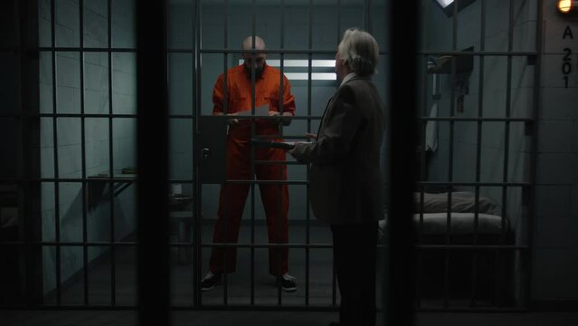 Gangster in orange uniform stands behind bars in prison cell, talks with advocate, reads lawyer contract. Inmate serves imprisonment term for crime in jail. Detention center or correctional facility.