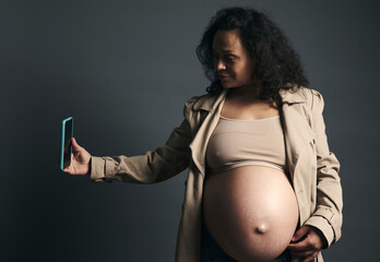 Attractive beautiful young gravid female, expectant mother photographing her tummy in third...