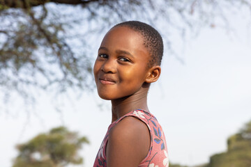 african girl with a big toothy smile , in the village, standing in the yard next to a tree