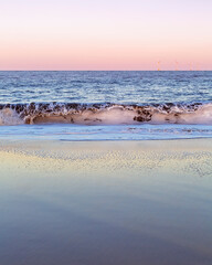 Gentle wave crashing on shimmering sandy beach with pink sunset sky, portrait orientation