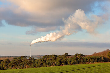 Tall slim industrial chimney billowing smoke into the sky in the countryside at golden hour
