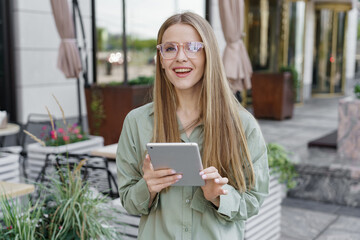 Smiling woman using a tablet outside a café, with a smart casual style and a bright urban setting.