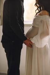 Pregnant woman in white dress and her husband standing near window and holding their hands on pregnant belly. Love concept