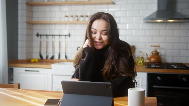 Smiling young asian woman sit in the kitchen having video call conversation through tablet computer. Share news, talk to family, enjoy webchat virtual meeting