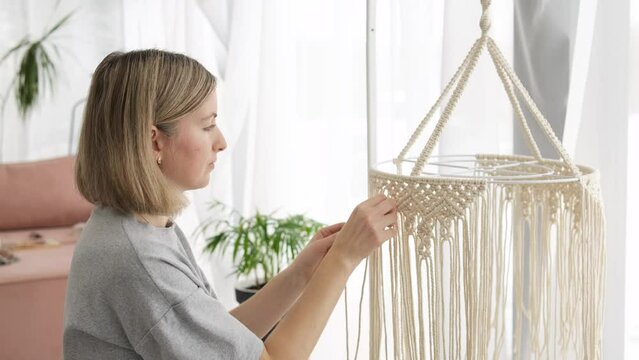 Woman making macrame lampshade with white threads at home. Woman knits ornament for lamp shade. Home hobby. Macrame knitting concept.