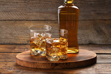 Whiskey with ice cubes in glasses and bottle on wooden table, closeup