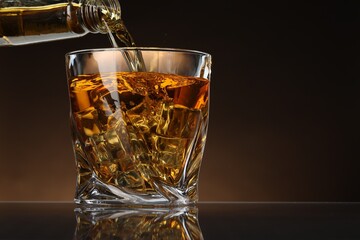 Pouring whiskey from bottle into glass with ice cubes at table against brown background, space for text